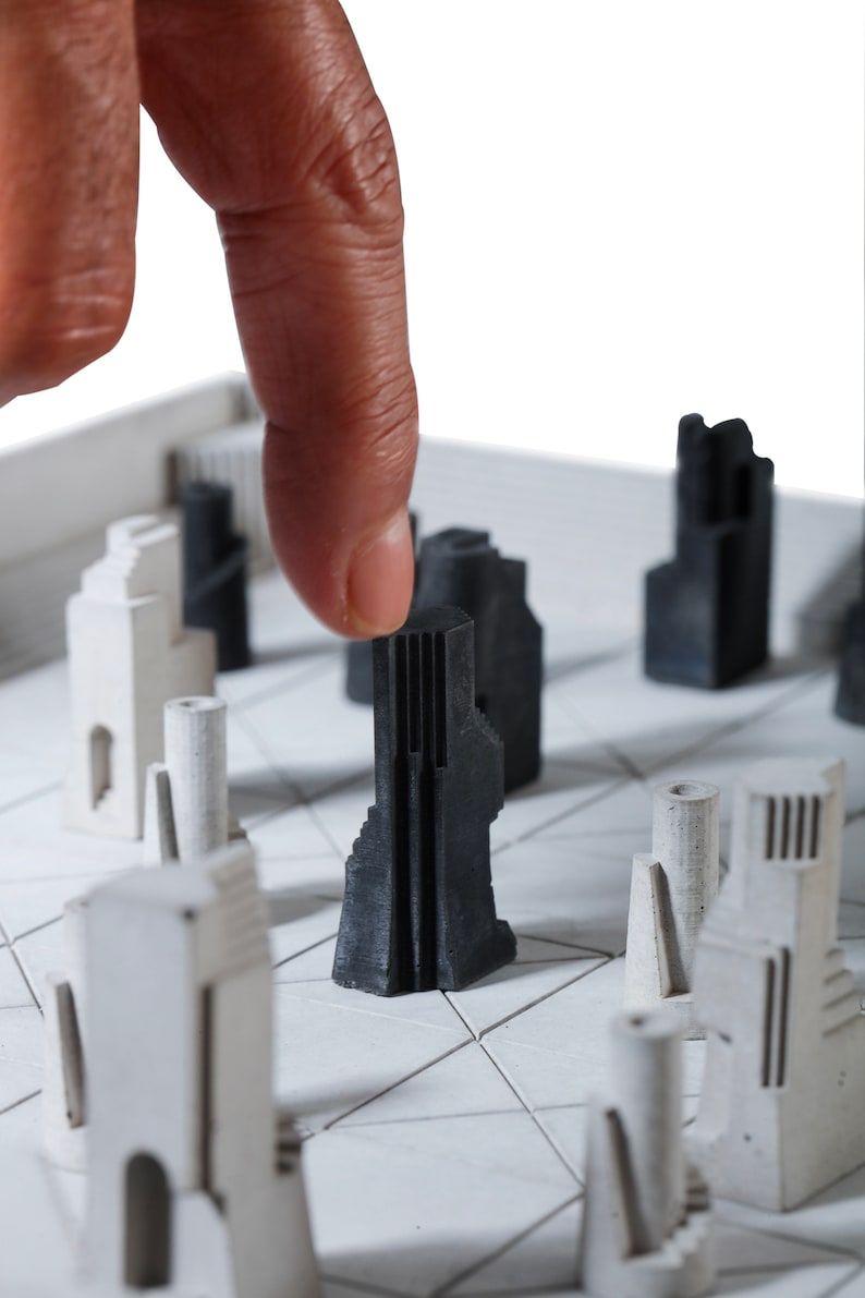 May include: A modern chess set featuring architectural-style pieces in black and white. The pieces are arranged on a white board with a grid pattern. A finger is touching a black piece, highlighting the game's strategic nature.