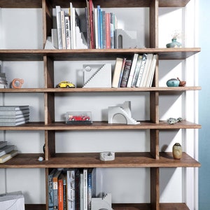 Puede incluir: Una estanter&iacute;a de madera con varios estantes, llena de libros y objetos decorativos. Los estantes son de madera oscura y tienen un fondo de madera clara. Hay varias peque&ntilde;as esculturas de hormig&oacute;n en los estantes, incluyendo algunas que se asemejan a escaleras. Tambi&eacute;n hay algunos coches de juguete peque&ntilde;os en los estantes.
