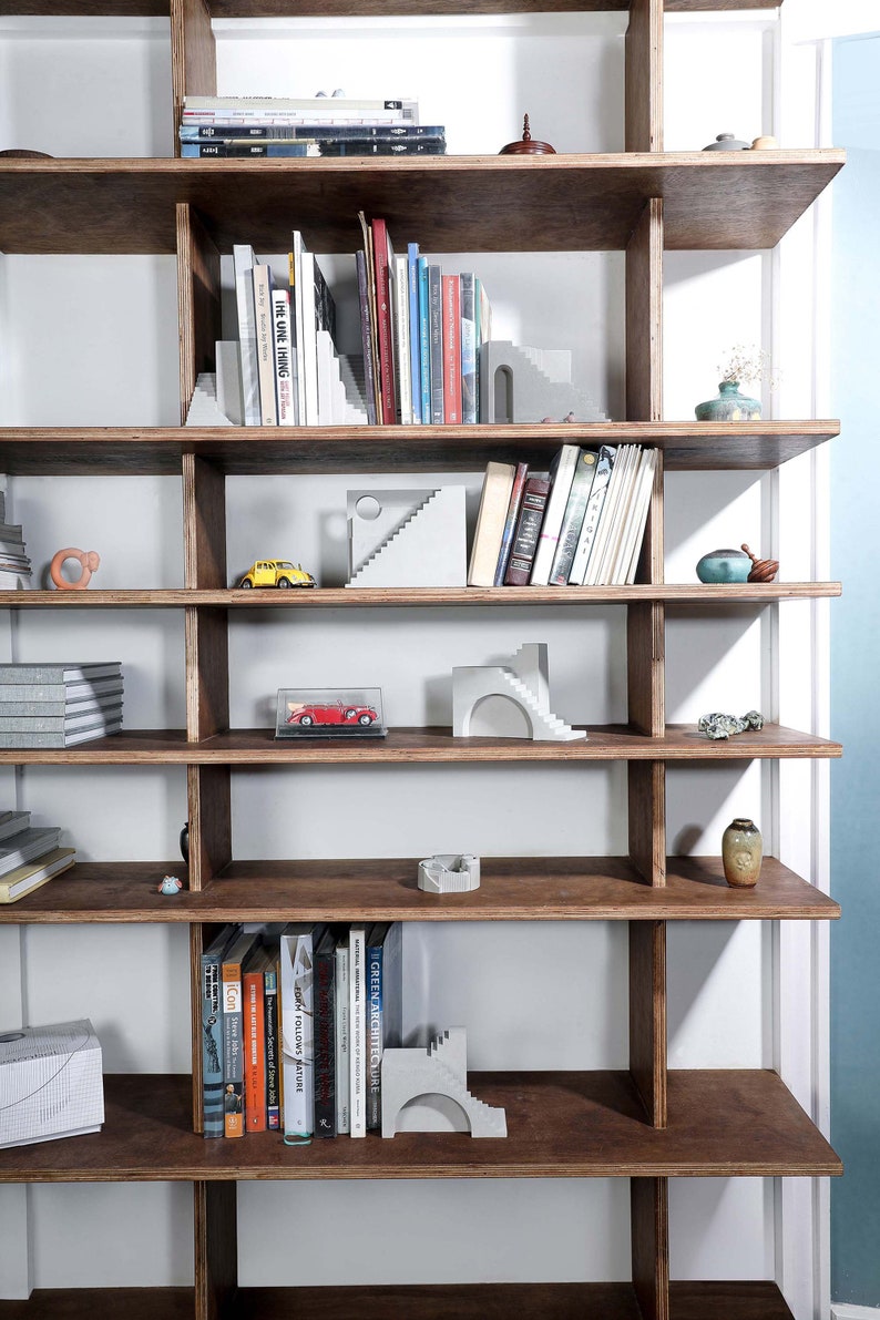 Puede incluir: Una estanter&iacute;a de madera con varios estantes, llena de libros y objetos decorativos. Los estantes son de madera oscura y tienen un fondo de madera clara. Hay varias peque&ntilde;as esculturas de hormig&oacute;n en los estantes, incluyendo algunas que se asemejan a escaleras. Tambi&eacute;n hay algunos coches de juguete peque&ntilde;os en los estantes.