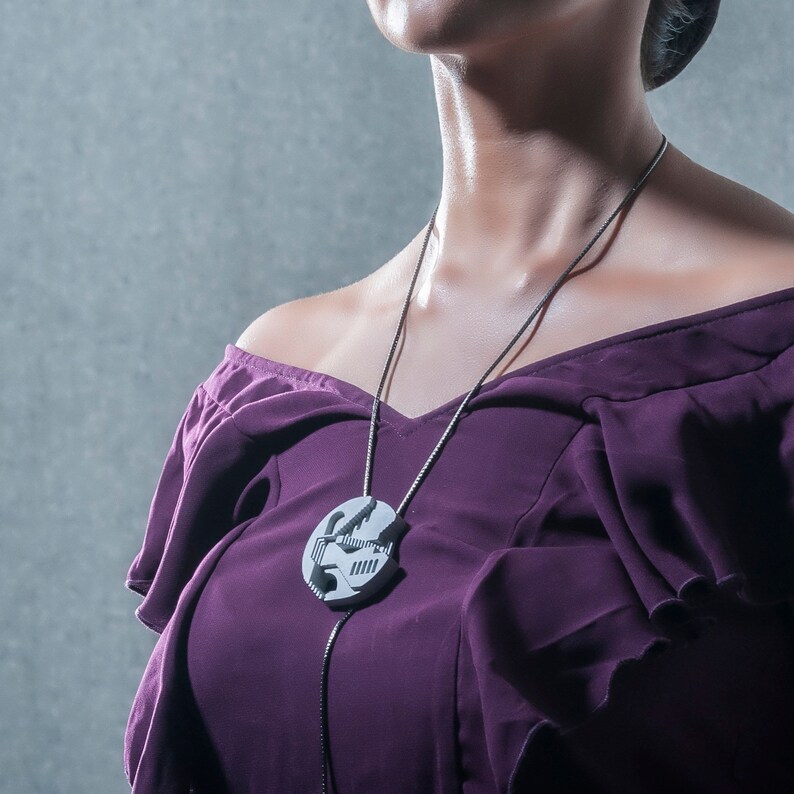 May include: A woman wearing a purple off-the-shoulder dress with a silver chain necklace featuring a geometric pendant.