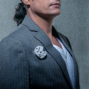 May include: A gray blazer with a white shirt underneath. A silver geometric pin is on the lapel of the blazer.