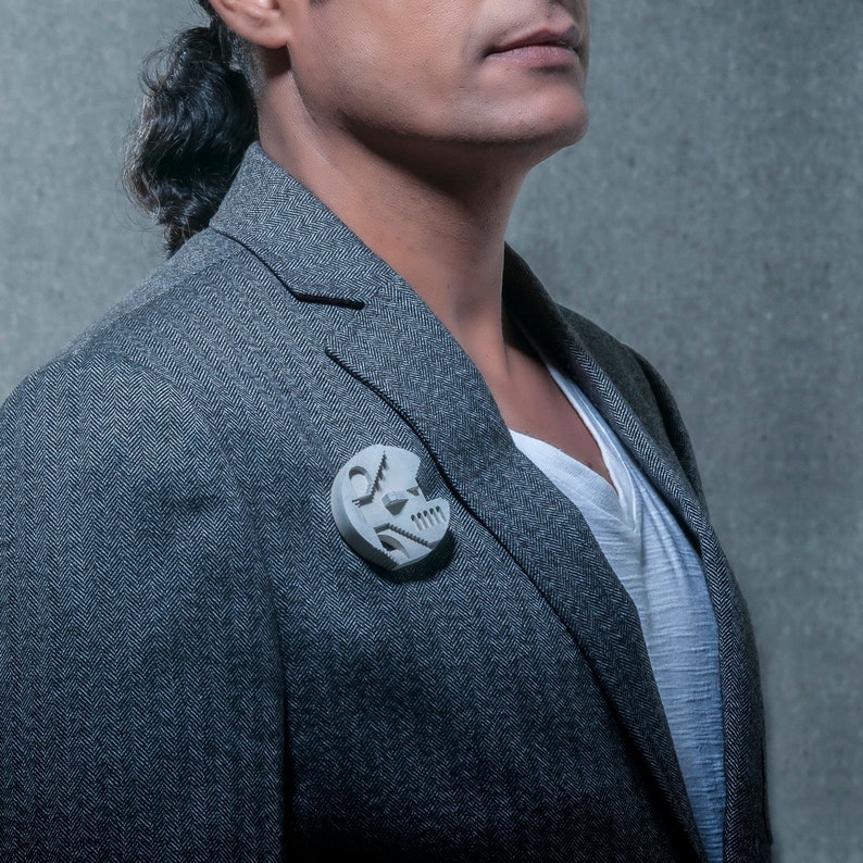 May include: A gray blazer with a white shirt underneath. A silver geometric pin is on the lapel of the blazer.