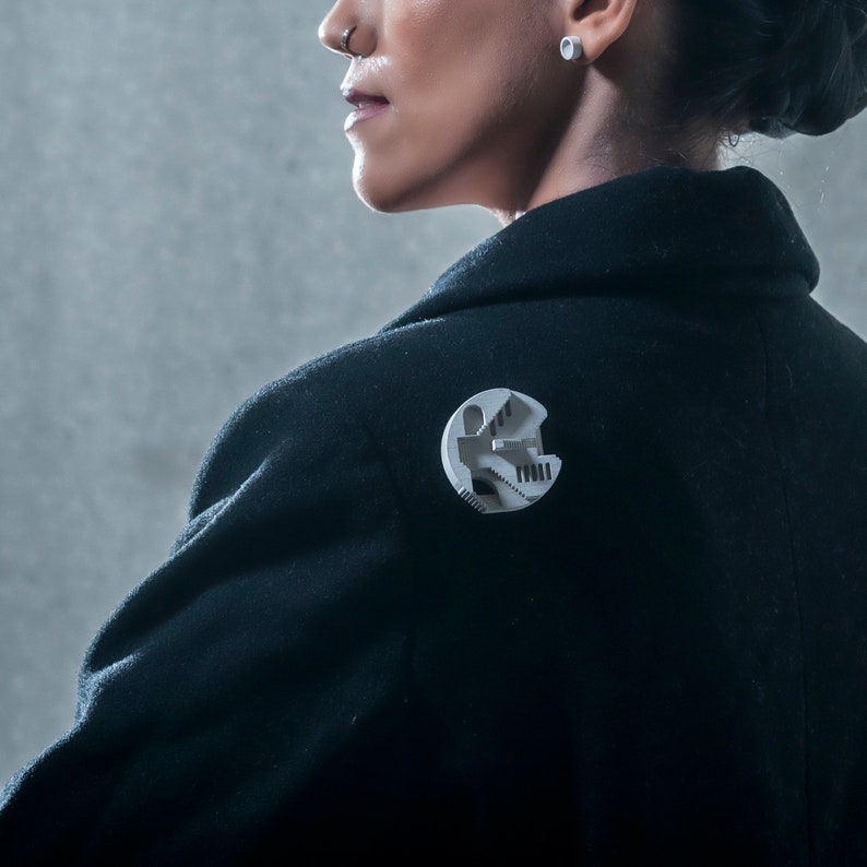 May include: A black blazer with a silver pin featuring a 3D design of a building with a staircase.