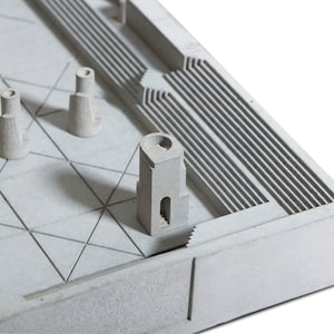 May include: A miniature architectural model in shades of gray, featuring various building forms and geometric patterns. The model includes a grid-like base, stepped elements, and tower-like structures, all rendered in a minimalist style.