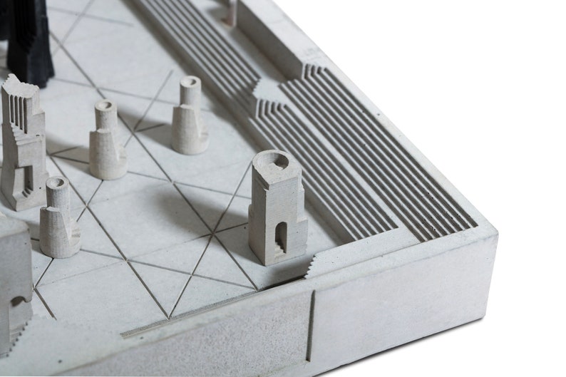 May include: A miniature architectural model in shades of gray, featuring various building forms and geometric patterns. The model includes a grid-like base, stepped elements, and tower-like structures, all rendered in a minimalist style.