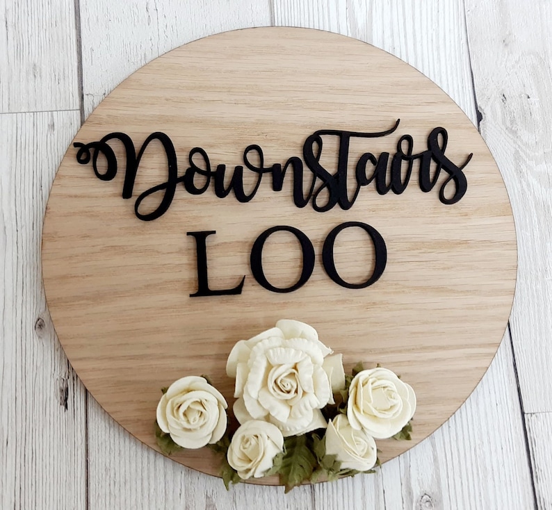 Pu&ograve; includere: Un cartello rotondo in legno con il testo "Downstairs Loo" in lettere nere. Il cartello &egrave; decorato con fiori di carta bianca.