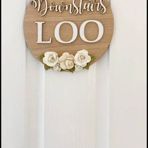Pu&ograve; includere: Un cartello rotondo in legno con le parole "Downstairs Loo" in lettere bianche. Il cartello &egrave; decorato con fiori bianchi.