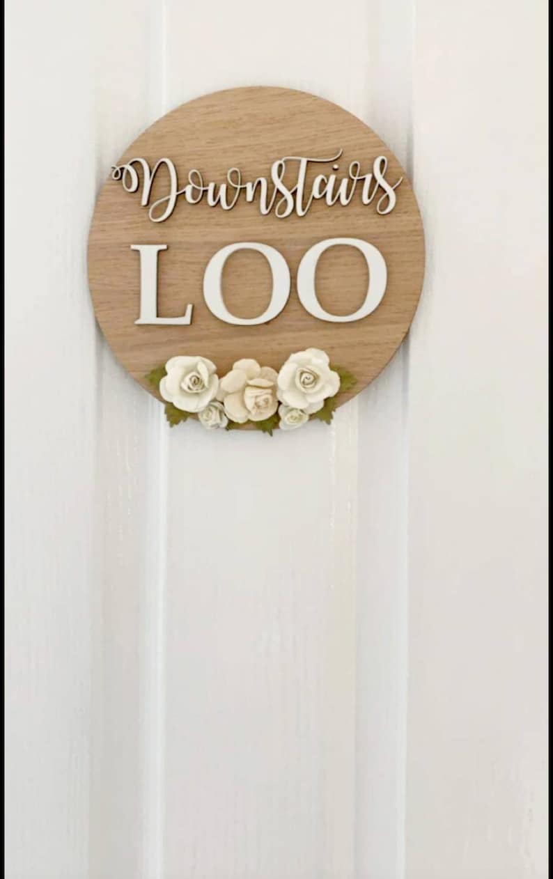 Pu&ograve; includere: Un cartello rotondo in legno con le parole "Downstairs Loo" in lettere bianche. Il cartello &egrave; decorato con fiori bianchi.