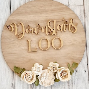 Pu&ograve; includere: Un cartello rotondo in legno con le parole "Downstairs Loo" in lettere dorate. Il cartello &egrave; decorato con un bordo di fiori bianchi.