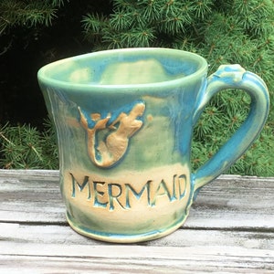 May include: A green and blue ceramic mug with a mermaid design and the word "MERMAID" on the front.