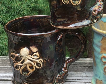 May include: Three ceramic mugs with octopus designs. The mugs are in different colors and styles. One mug is brown with a white octopus, one is black with a white octopus, and one is blue with a gold octopus.