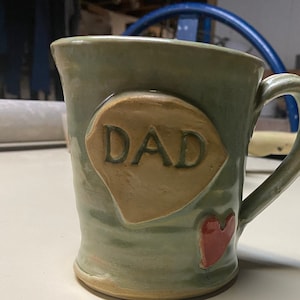 Puede incluir: Taza de cerámica hecha a mano con un esmalte verde claro. La taza presenta un adorno en forma de corazón beige con la palabra "DAD" y un pequeño corazón rojo. La taza tiene un asa curvada.