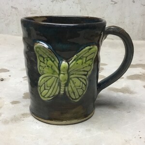 May include: A handmade ceramic mug with a dark blue-green glaze and a raised, light green butterfly design. The mug has a rounded handle and a textured surface, perfect for enjoying coffee or tea.