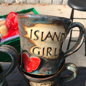 Puede incluir: Dos tazas de cerámica con asas. La taza de arriba es azul, verde y marrón con un corazón rojo y las palabras "Island Girl". La taza de abajo es marrón con las palabras "Beach Girl".
