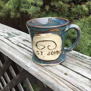 May include: A blue and brown ceramic mug with a white oval design featuring a swirl pattern and the text "ST JOHN".