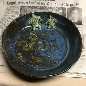 May include: A blue ceramic bowl with a decorative design of small circles and two green turtle figurines. The bowl is sitting on a newspaper with the headline "Couple stages festival for 2 sons lost to guns".