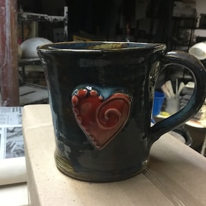 Puede incluir: Una taza de cerámica azul oscuro con un diseño de corazón rojo. El corazón tiene un patrón de espiral y está rodeado de puntos blancos.