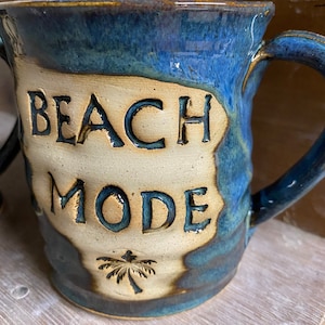 Può includere: Una tazza in ceramica blu e marrone con le parole "Beach Mode" e un disegno di palma.