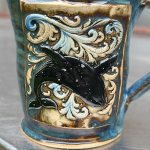 May include: A ceramic mug with a glossy blue and brown glaze featuring a raised, black orca whale relief centered within an intricate, cream-colored scrollwork design. The mug has a sturdy handle and a textured, multi-toned finish throughout.