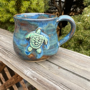 May include: A blue and green ceramic mug with a raised sea turtle design. The mug has a brown glaze and a speckled finish.