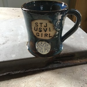 Op de afbeelding: Een handgemaakte keramische mok met een donkerblauwe glazuur en een bruine basis. De mok heeft de tekst "STJ USVI GIRL" en een wit zeesterontwerp. De handgreep is gebogen.