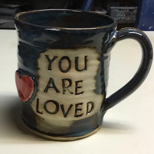 Op de afbeelding: Handgemaakte keramische mok met een donkerblauwe glazuur en een crèmekleurig midden. De mok heeft de woorden "YOU ARE LOVED" en een rood hart. Het handvat is donkerblauw.