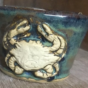 May include: A blue and brown ceramic planter with a white crab design. The planter has a glazed finish and a rounded shape.