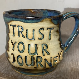 Op de afbeelding: Een keramische mok met een lichtbruine glazuur en blauwe accenten. Op de mok staat "TRUST YOUR JOURNEY" in donkerblauwe glazuur.
