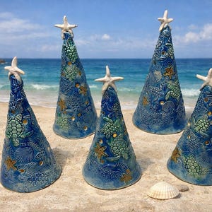 May include: Six ceramic cone-shaped sculptures with a blue glaze, decorated with sea turtles, shells, and starfish. Each sculpture is topped with a white starfish. The sculptures are on a sandy beach with the ocean in the background.