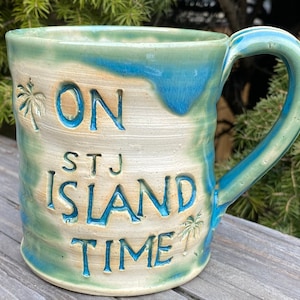 May include: A blue and green ceramic mug with the text "ON STJ ISLAND TIME" and palm tree designs.