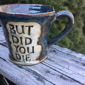 May include: A blue and brown ceramic mug with a white inscription that reads "BUT DID YOU DIE".