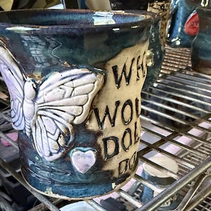 May include: A ceramic mug with a dark blue and green glaze featuring a raised, light-colored butterfly and a small heart. The mug has embossed text that reads "WHO WOULD DO THAT" in black lettering on a cream-colored panel on the side.
