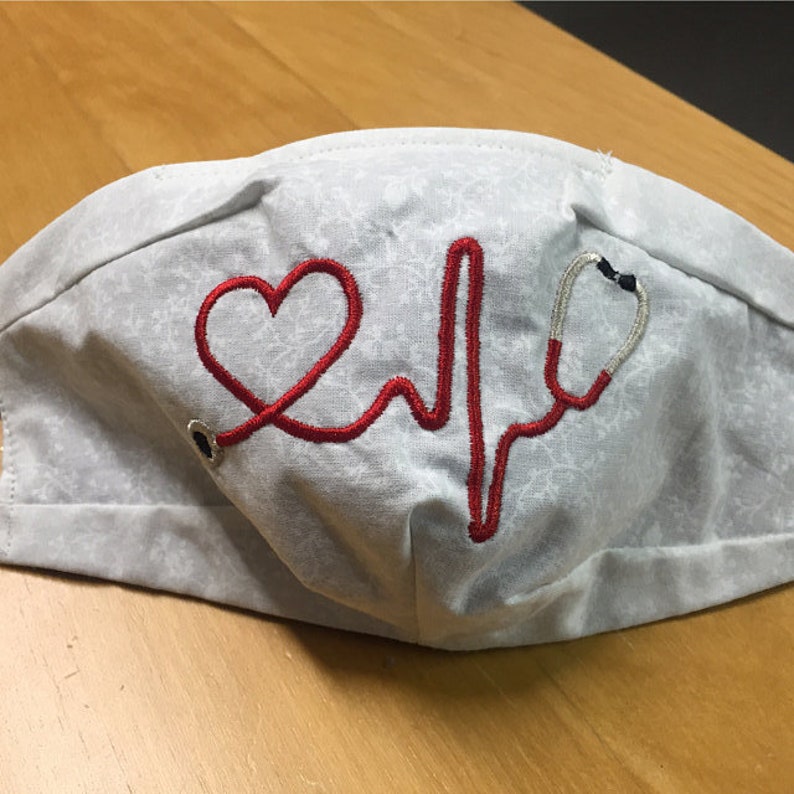 May include: A white face mask with a red embroidered heart and a stethoscope with a heartbeat line. The mask has a subtle gray floral pattern.