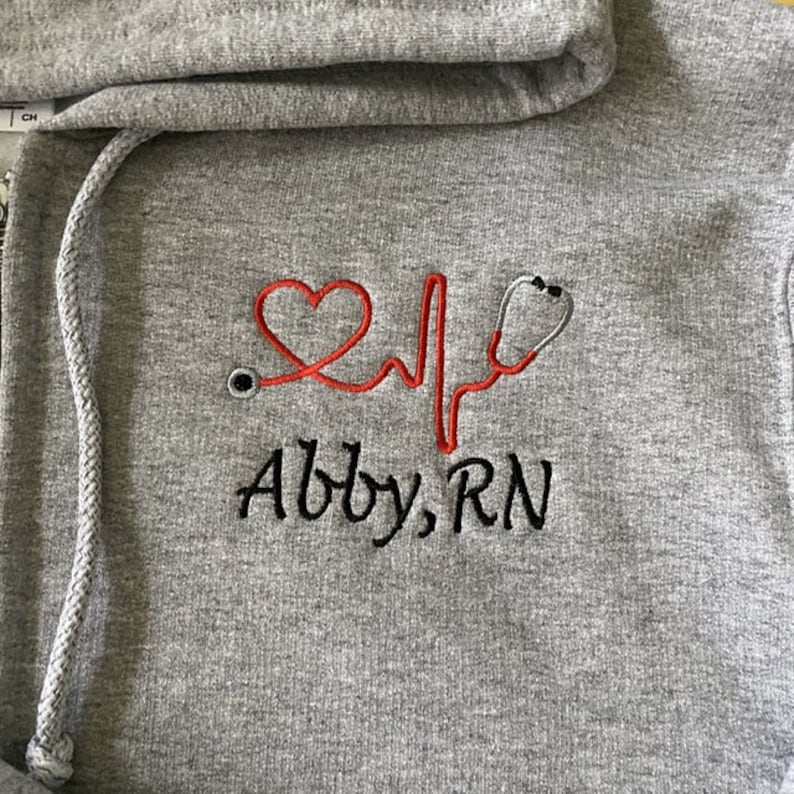 May include: Gray hooded sweatshirt with red embroidered heart, stethoscope, and heartbeat line. The text "Abby, RN" is embroidered in black below the design.