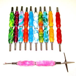 May include: A collection of colorful seam rippers and a pink seam ripper with a pair of scissors. The seam rippers have marbled handles in various colors, including green, red, blue, orange, and yellow. The tools are arranged on a white background.