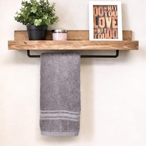 Op de afbeelding: Een houten plank met een zwarte handdoekstang, met een grijze handdoek. De plank toont ook een ingelijste print met de woorden "DO WHAT YOU LOVE" en een kleine potplant en een kaars.