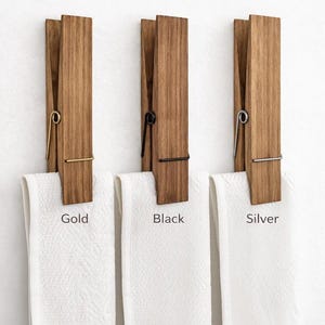 May include: Wooden clothespin-style towel holders with white towels. The holders have gold, black, and silver metal accents. The text "CHOOSE YOUR FINISH" is at the top, with the words "Gold", "Black", and "Silver" below each holder.