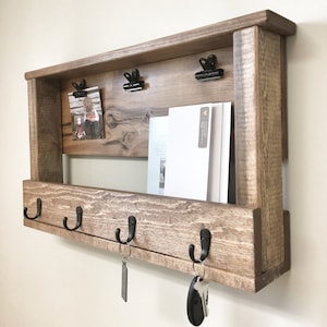 May include: Rustic wooden wall organizer with a shelf, hooks, and clips. The shelf has a picture and some papers on it. The hooks have keys attached to them.