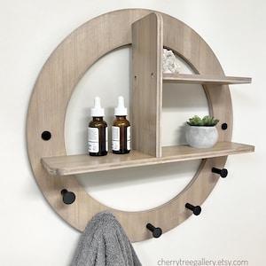 May include: A round wooden wall shelf with three hooks and two shelves. The shelf is made of light wood and has a natural finish. The shelves are decorated with small plants and bottles. The hooks are black metal and are attached to the bottom of the shelf. The shelf is mounted on a white wall. cherrytreegallery.etsy.com