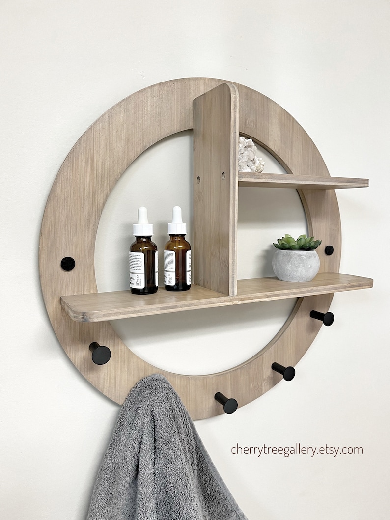 May include: A round wooden wall shelf with three hooks and two shelves. The shelf is made of light wood and has a natural finish. The shelves are decorated with small plants and bottles. The hooks are black metal and are attached to the bottom of the shelf. The shelf is mounted on a white wall. cherrytreegallery.etsy.com