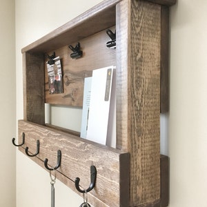 May include: A rustic wooden wall organizer with a shelf, hooks, and a memo board. The organizer is stained in a dark brown color and has a distressed finish. It features three hooks for hanging keys and a shelf for storing mail or other items. The memo board has two clips for holding notes or photos.