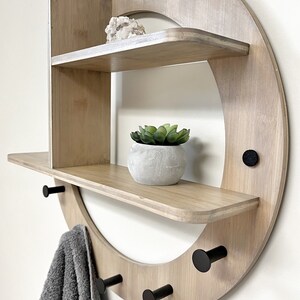 May include: A round wooden wall shelf with two shelves and four black hooks. The shelf has a light wood finish and is decorated with a small potted plant and a crystal.