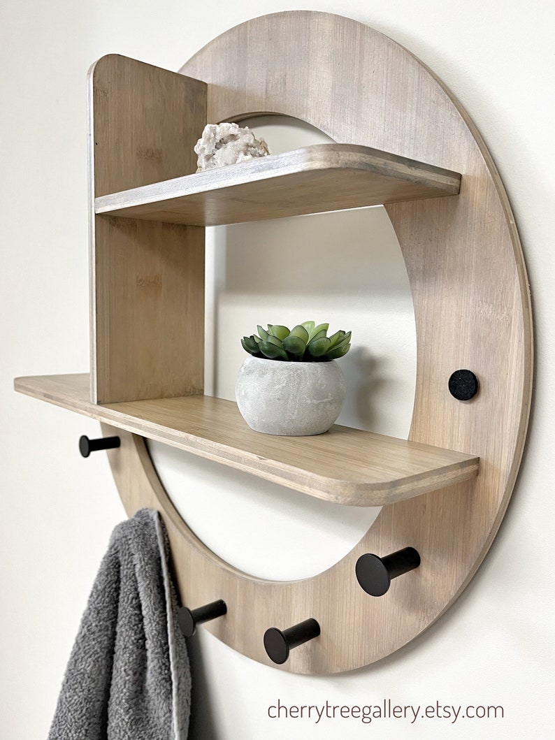 May include: A round wooden wall shelf with two shelves and four black hooks. The shelf has a light wood finish and is decorated with a small potted plant and a crystal.