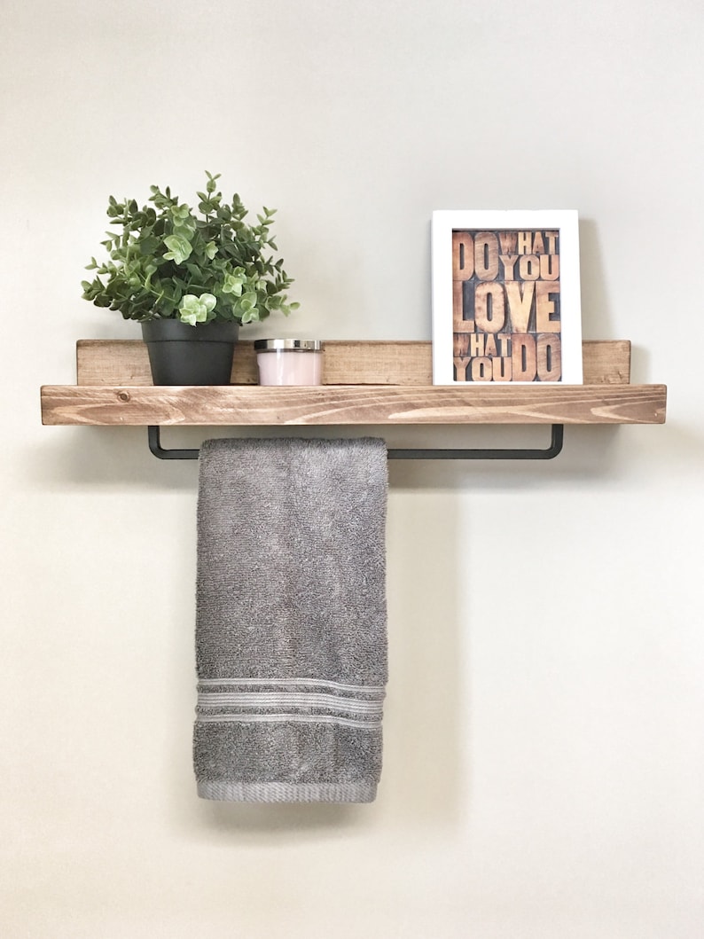 Op de afbeelding: Een houten plank met een zwarte handdoekstang, met een grijze handdoek. Een kleine potplant, een kaars en een ingelijste print met de woorden "DO WHAT YOU LOVE" staan op de plank. De plank is aan een lichtgekleurde muur bevestigd.