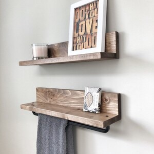 May include: Two rustic wooden shelves mounted on a white wall. The top shelf holds a framed print with the text "DO WHAT YOU LOVE WHAT YOU DO" and a candle. The bottom shelf has a towel bar and a gray towel.