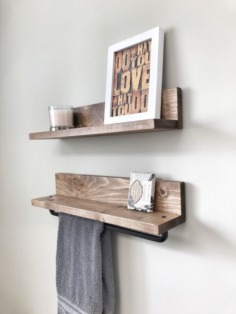 May include: Two rustic wooden shelves mounted on a white wall. The top shelf holds a framed print with the text "DO WHAT YOU LOVE WHAT YOU DO" and a candle. The bottom shelf has a towel bar and a gray towel.