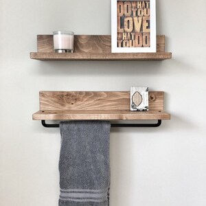 May include: Two wooden floating shelves mounted on a light gray wall. The top shelf holds a candle and a framed print that reads "DO WHAT YOU LOVE WHAT YOU DO." The bottom shelf has a towel bar with a gray towel.