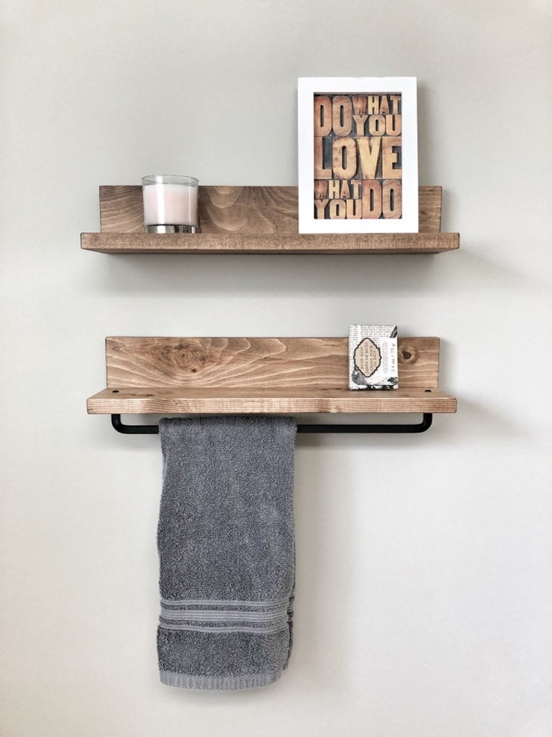 May include: Two wooden floating shelves mounted on a light gray wall. The top shelf holds a candle and a framed print that reads "DO WHAT YOU LOVE WHAT YOU DO." The bottom shelf has a towel bar with a gray towel.