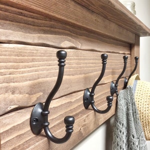 May include: A rustic wooden wall-mounted coat rack with four black metal hooks. The rack has a shelf on top and is made of stained wood. A gray sweater and a straw hat hang from the hooks.