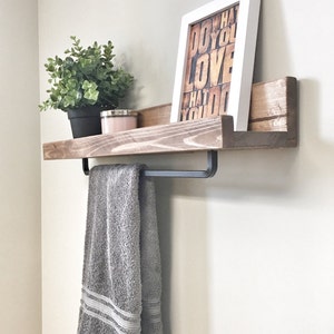 Op de afbeelding: Een houten plank met een handdoekenrek, met een grijze handdoek, een ingelijste print met de tekst "DO WHAT YOU LOVE", een kleine potplant en een kaars. De plank is aan een lichtgekleurde muur bevestigd.